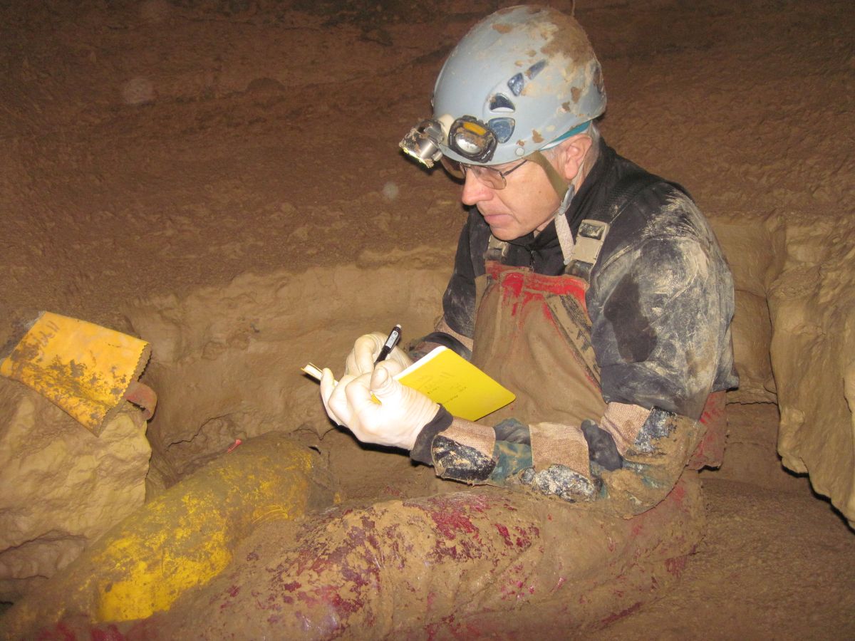 A caver in a helmet and headlamp sketches in notepad while sitting in a narrow, underground passage, mud caking his clothing.