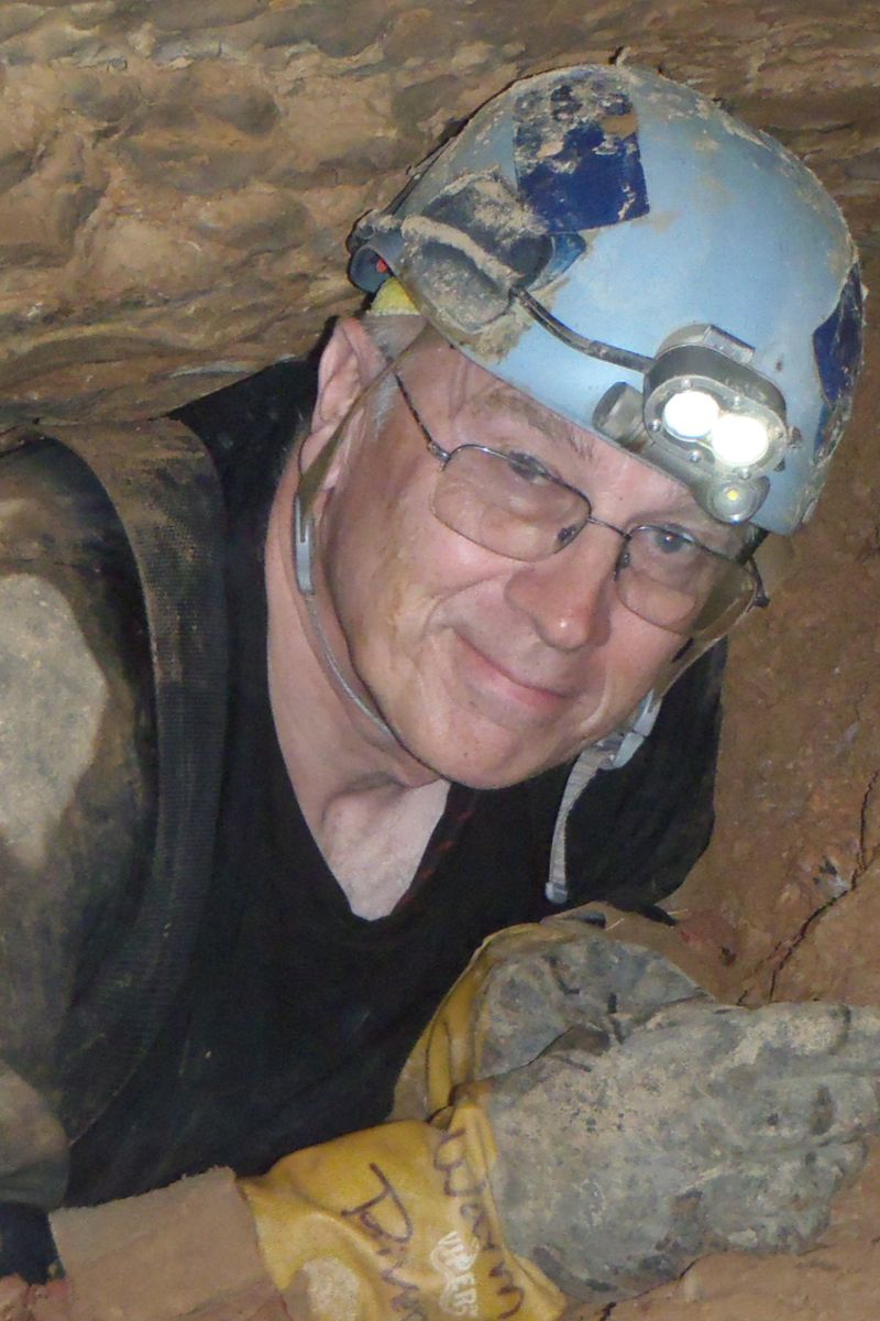 Smiling man wearing glasses and a muddy blue helmet with a headlamp inside a cave.
