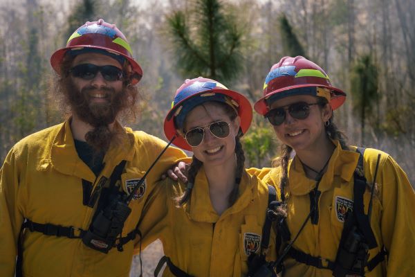 Three people dressed in protective gear in front of prescribed burn.